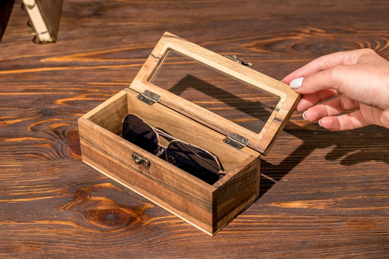 Puede incluir: Una caja de madera con tapa de vidrio, abierta para revelar un par de gafas de sol en su interior. La caja est&aacute; hecha de madera marr&oacute;n claro con un acabado natural. Las gafas de sol son negras con un marco dorado.
