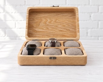 May include: A rectangular light-colored wooden watch box with a hinged lid and a metal latch. The interior features six individual compartments, each containing a beige fabric-covered cushion. Two watches are displayed on the cushions.