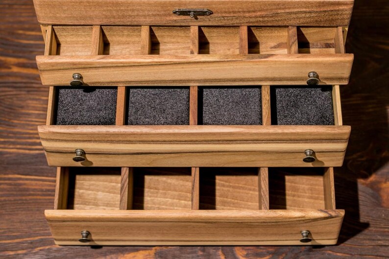 May include: A wooden jewelry box with three drawers. The top drawer has multiple compartments, the middle drawer has four compartments lined with black felt, and the bottom drawer has multiple compartments.