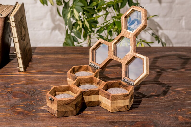 K&ouml;nnte beinhalten: Eine sechseckige Holzschachtel f&uuml;r Ringe mit einem Scharnierdeckel. Die Schachtel ist aus hellem und dunklem Holz gefertigt und hat f&uuml;nf F&auml;cher, die mit grauem Filz ausgekleidet sind. Der Deckel ist ge&ouml;ffnet und zeigt eine transparente Glasscheibe.