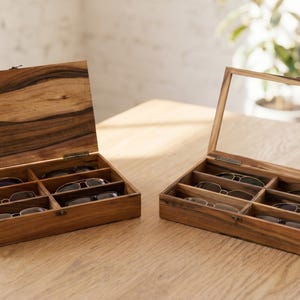 以下が含まれることがあります： 明るい木製の表面に、2つの木製メガネケースが置かれています。1つのケースはヒンジ付きの蓋が開いており、6つのコンパートメントにメガネが入っています。もう1つのケースは透明なガラスの蓋が付いています。どちらのケースも、自然な木目を持つ濃い色の木材で作られています。