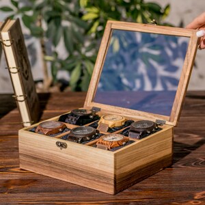 May include: A rectangular wooden watch box with a hinged glass lid, featuring six individual compartments lined with dark fabric. Each compartment contains a wooden wristwatch with a round face and a matching wooden link strap in various brown tones.