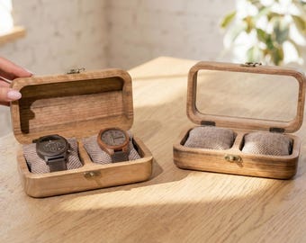 May include: Two wooden watch display boxes. One box is open, revealing two watches with dark faces and leather straps. The other box is closed with a clear glass top. Both boxes are on a wooden surface.