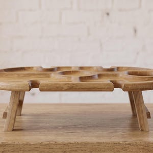 Könnte beinhalten: Ein hellbrauner Picknicktisch aus Holz mit niedrigem Profil und vier kurzen Beinen. Die Tischplatte verfügt über eine mittige kreisförmige Vertiefung, zwei seitliche kreisförmige Aussparungen und vier schmale Schlitze an den Rändern für Gläser.
