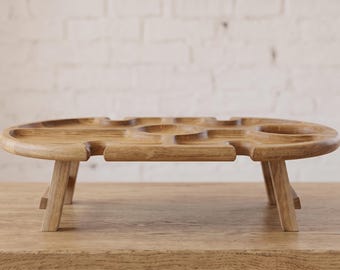 May include: A light brown wooden picnic table with a low profile and four short legs. The tabletop features a central circular indentation, two side circular cutouts, and four narrow slots along the edges for holding glasses or stemware securely.
