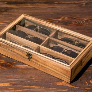 Puede incluir: Una caja de madera con tapa de vidrio para guardar gafas de sol. La caja tiene seis compartimentos y est&aacute; hecha de madera oscura con veta natural. La caja est&aacute; sobre una superficie de madera.