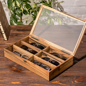 Puede incluir: Una caja de madera con tapa de vidrio, que contiene seis pares de gafas de sol. La caja tiene un acabado de madera marr&oacute;n claro y est&aacute; abierta, revelando las gafas de sol en el interior.