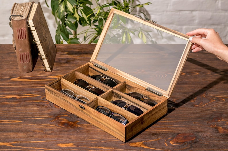 Puede incluir: Una caja de madera con tapa de vidrio, que contiene seis pares de gafas de sol. La caja tiene un acabado de madera marr&oacute;n claro y est&aacute; abierta, revelando las gafas de sol en el interior.