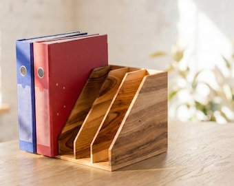 May include: A wooden desk organizer with four angled compartments and two binders. The binders are red and blue. The organizer is made of a light-colored wood with a natural grain. The background is blurred, with a window and plants visible.