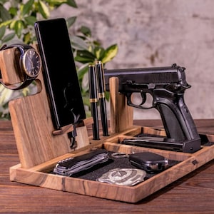 May include: Wooden desk organizer with compartments for a watch, smartphone, pens, a handgun, a knife, keys, and a car key fob. The organizer is made of dark wood and has a black felt lining in the compartments.