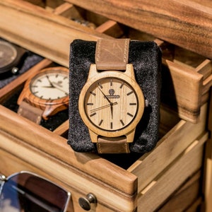May include: A wooden watch with a brown leather band in a wooden box with other watches. The watch face is light wood with black hands and a small logo that says "Sunwood".