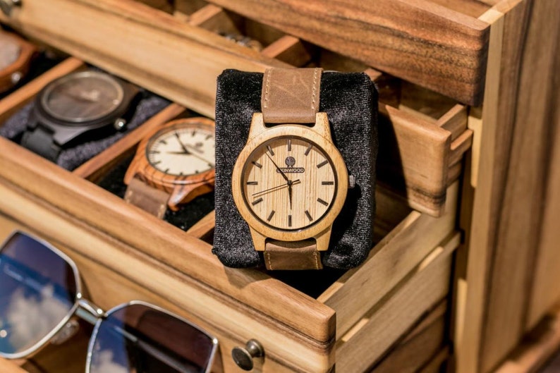May include: A wooden watch with a brown leather band in a wooden box with other watches. The watch face is light wood with black hands and a small logo that says "Sunwood".