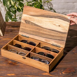 Puede incluir: Una caja de madera con tapa con bisagras, abierta para revelar seis compartimentos para guardar gafas. La caja est&aacute; hecha de madera marr&oacute;n claro con un acabado de veta de madera natural.