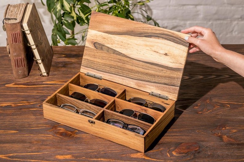 Puede incluir: Una caja de madera con tapa con bisagras, abierta para revelar seis compartimentos para guardar gafas. La caja est&aacute; hecha de madera marr&oacute;n claro con un acabado de veta de madera natural.