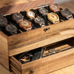 Op de afbeelding: Een rechthoekige houten horlogedoos met een scharnierend deksel en een uitschuifbare lade. Het bovenste compartiment heeft acht vakken met zwarte kussentjes, elk met een houten horloge. De onderste lade bevat diverse kleine metalen accessoires en munten.