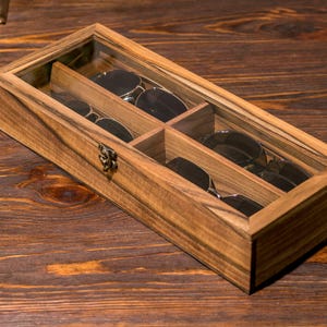 Puede incluir: Una caja de madera con tapa de vidrio para guardar gafas de sol. La caja tiene cuatro compartimentos y est&aacute; hecha de madera oscura con un acabado natural.