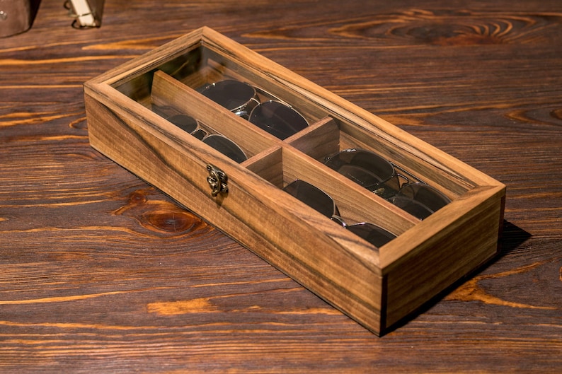 Puede incluir: Una caja de madera con tapa de vidrio para guardar gafas de sol. La caja tiene cuatro compartimentos y est&aacute; hecha de madera oscura con un acabado natural.