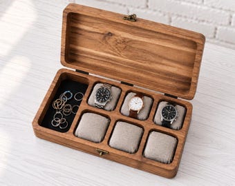 May include: A rectangular wooden watch box with a hinged lid and a brass latch. The interior features a black velvet compartment for rings and five individual sections with beige fabric cushions, three of which hold wristwatches.