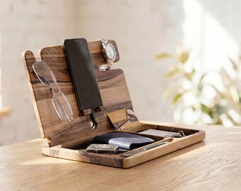 May include: Wooden desk organizer with compartments for a smartphone, glasses, watch, wallet, and other small items. The organizer is made of a light brown wood and sits on a wooden surface. The design includes a slot for the phone and a tray for accessories.