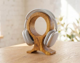 May include: A wooden headphone stand with a circular design, holding a pair of silver headphones. The stand is made of light brown wood and sits on a wooden surface. The headphones are a light silver color.