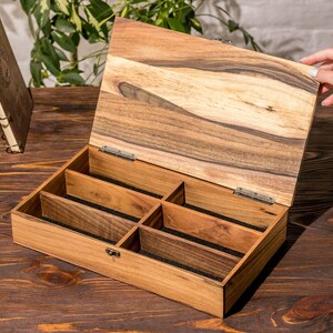 Puede incluir: Una caja de madera con tapa con bisagras y seis compartimentos forrados con fieltro negro. La caja est&aacute; hecha de madera marr&oacute;n claro con veta de madera marr&oacute;n oscuro.