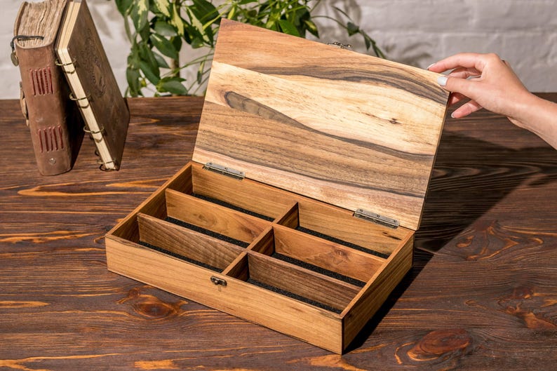 Puede incluir: Una caja de madera con tapa con bisagras y seis compartimentos forrados con fieltro negro. La caja est&aacute; hecha de madera marr&oacute;n claro con veta de madera marr&oacute;n oscuro.