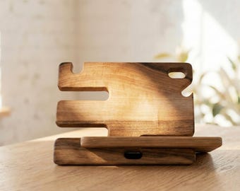 May include: Wooden phone stand with a slot for a charging cable. The stand is made of light brown wood and has a unique design with multiple cutouts. It is sitting on a wooden table, with a window and plants in the background.