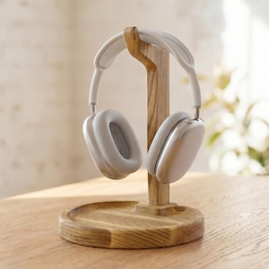 May include: White over-ear headphones on a wooden stand. The stand is made of light-coloured wood and has a circular base. The headphones are white with grey earcups. The stand is on a wooden table. The headphones are a modern design.