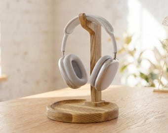 May include: A pair of white over-ear headphones rests on a wooden headphone stand. The stand is made of light-colored wood and has a circular base. The headphones are white with gray earcups. The stand is on a wooden table.