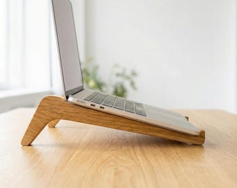May include: A wooden laptop stand with a natural finish. The stand is angled to elevate a laptop, improving ergonomics. The laptop is silver and black. The stand is on a light-colored wooden surface.