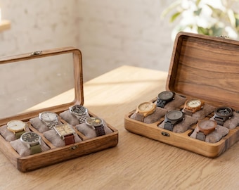 May include: Two wooden watch display cases. One has a clear lid, the other is open. Each case holds several watches on small, tan cushions. The cases are made of dark wood and sit on a light-colored wooden surface.