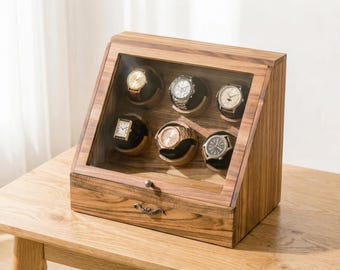 May include: A wooden watch display case with a glass front, showcasing six wristwatches. The case is made of light brown wood and has a drawer. The watches have various band and face designs, including gold, silver, and black.