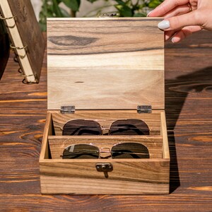Puede incluir: Una caja de madera con tapa con bisagras, abierta para revelar dos pares de gafas de sol. La caja es de madera oscura con un patr&oacute;n de veta natural. Las gafas de sol son marrones y tienen un estilo aviador cl&aacute;sico.