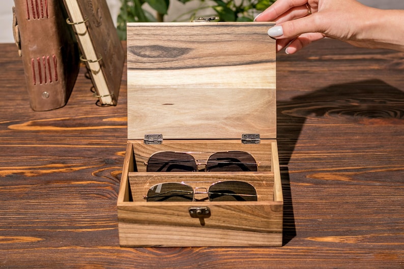 Puede incluir: Una caja de madera con tapa con bisagras, abierta para revelar dos pares de gafas de sol. La caja es de madera oscura con un patr&oacute;n de veta natural. Las gafas de sol son marrones y tienen un estilo aviador cl&aacute;sico.