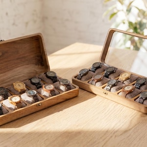 May include: Two wooden watch display cases, one open and one closed, each holding multiple wristwatches. The cases are made of light-colored wood and have a clear glass top. The watches have various band and face colors.