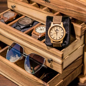 May include: Wooden box with multiple drawers for storing watches and sunglasses. The top drawer has a black velvet lining and contains a wooden watch with a brown leather band. The bottom drawer contains a pair of sunglasses.