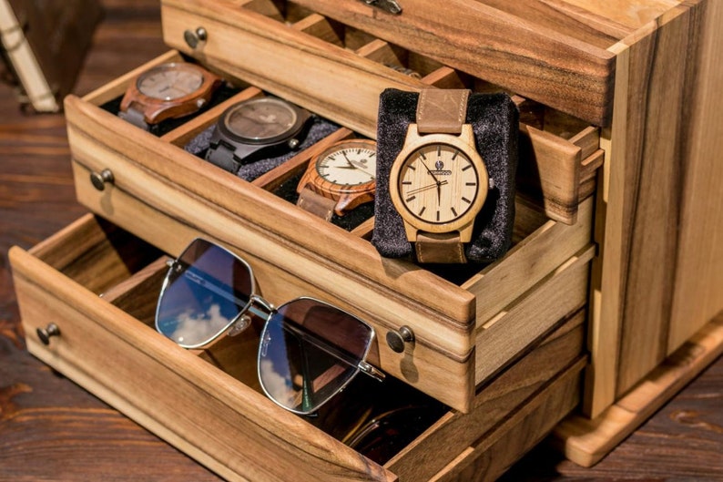 May include: Wooden box with multiple drawers for storing watches and sunglasses. The top drawer has a black velvet lining and contains a wooden watch with a brown leather band. The bottom drawer contains a pair of sunglasses.