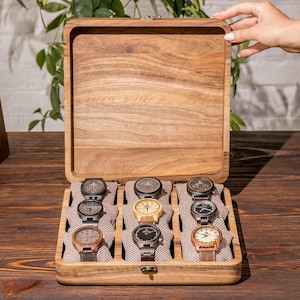 Puede incluir: Una caja de madera para relojes con una tapa con bisagras, abierta para revelar nueve compartimentos para relojes. Los compartimentos están forrados con una tela suave de color marrón y cada uno contiene un reloj de pulsera con un diseño diferente.