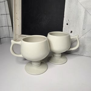 May include: Two off-white ceramic mugs with pedestal bases and curved handles. The mugs have a rounded bowl shape and are displayed on a white surface. The background includes a white framed chalkboard and a white wooden panel.