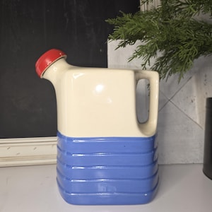 May include: A vintage ceramic pitcher with a red lid and handle. The top half is cream-colored, while the bottom half features horizontal blue stripes. The pitcher is designed with a spout and a handle for easy pouring.