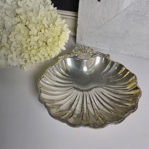 May include: A silver-toned metal dish shaped like a scallop shell. The dish features a fluted, radiating pattern that extends from the center to the scalloped edges, with a decorative shell-like motif at the top edge and visible tarnish.