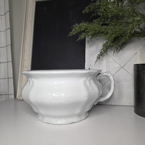 May include: A white, antique-style chamber pot with a handle. The ceramic pot has a fluted design and a wide, rounded shape. The pot is sitting on a white surface.
