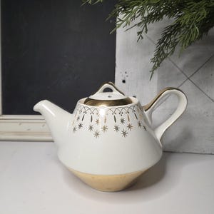 May include: A white ceramic teapot with gold accents. The teapot features a gold rim, handle, and base. Delicate gold star and line patterns adorn the body. A small, gold-trimmed lid sits atop the teapot. The teapot is a vintage style.