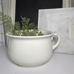 May include: A white ceramic chamber pot with a rounded body, a wide rim, and a single side handle. The vessel is filled with green fern fronds, showcasing a smooth, glossy finish and a classic, deep basin design suitable for various decorative uses.