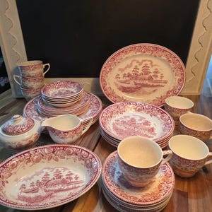 May include: A collection of ceramic dinnerware featuring a red and white transferware pattern of rural landscapes and floral borders. The set includes plates, bowls, cups, saucers, a lidded sugar bowl, and a creamer, all with matching designs.