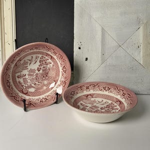May include: Two red and white Willow pattern ceramic bowls. One is a shallow plate, the other a deeper bowl. Both have a detailed Asian-inspired landscape design and a decorative border. The bowls are displayed on a white surface.