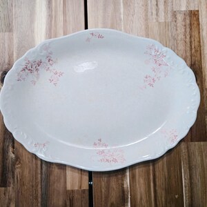May include: A white oval serving platter with a delicate pink floral pattern. The platter has a slightly raised rim and a scalloped edge.