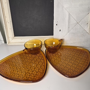 May include: Two amber-colored glass teacups with matching triangular plates. The plates have a textured, geometric pattern. The teacups have handles. The set is displayed on a white surface.