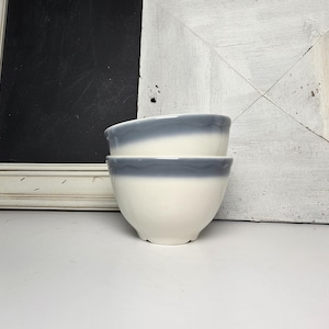 May include: Two white ceramic bowls with a blue-grey rim, stacked on top of each other. The bowls have a simple, rounded shape and are set against a white and black background.