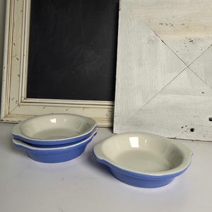 May include: Three oval ceramic dishes with a smooth, glossy finish. Each dish features a light blue exterior and a cream-colored interior, complete with small integrated handles on opposite sides for easy gripping and handling.